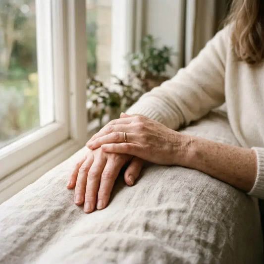 Close-up of a woman's hands resting gently on a soft linen-covered lap beside a sunlit window, showing natural skin texture and subtle age spots in warm morning light.