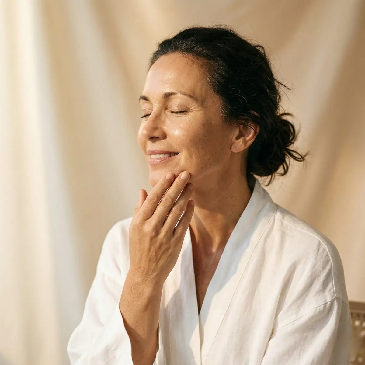 Mature woman in her 50s gently touching her jawline with eyes closed, glowing hydrated skin, wearing a white robe in soft warm light, menopausal skincare routine