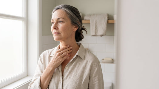 Woman in her fifties resting her fingertips at the base of her throat in soft daylight, thyroidectomy scar care after 45