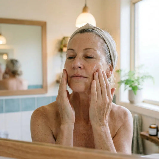  Woman in her 50s applying a clear hydrating serum to damp skin in front of a morning bathroom mirror