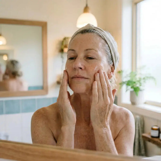  Woman in her 50s applying a clear hydrating serum to damp skin in front of a morning bathroom mirror