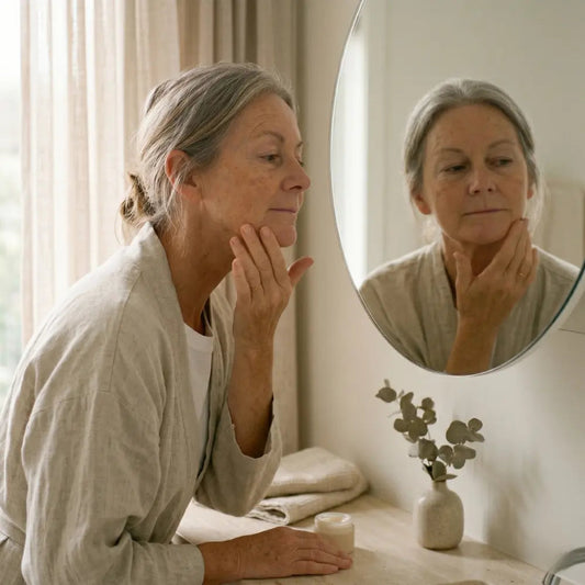 Australian woman in her early 50s gently massaging firming cream upward along her jawline at a bathroom vanity in soft natural morning light