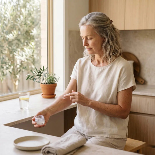 Australian woman in her 50s applying a small amount of scar gel to her forearm in soft natural light at a kitchen window