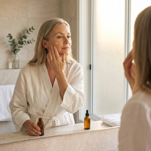 Woman in her 50s applying a pale yellow vitamin C serum to her cheek in soft morning light