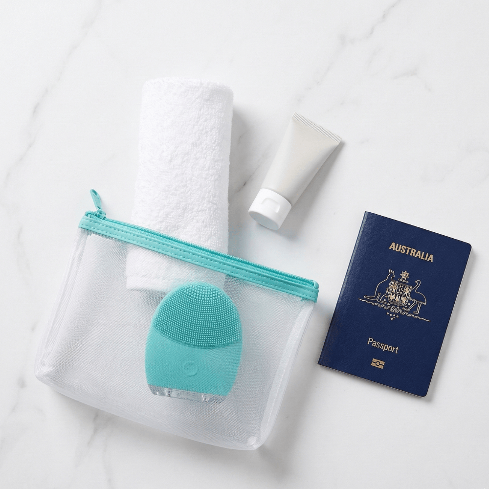 Genova GO Silicone Facial Cleanser in clear travel pouch with cleanser, towel and passport on marble, compact portable face cleansing device perfect for travel skincare routine